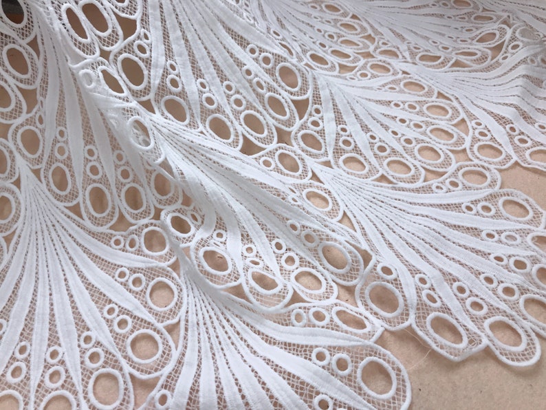 May include: White lace fabric with a detailed peacock feather design. The fabric features a repeating pattern of open circles and curved lines, creating a delicate and airy texture. Ideal for crafting or apparel.