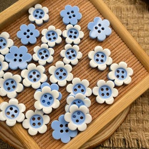 May include: A collection of flower-shaped buttons featuring a two-tone design with light blue centers and white outer petals. Each button has four holes in the center for sewing. The buttons are arranged in a scattered pattern on a flat surface.