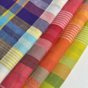May include: A collection of folded fabric swatches featuring various plaid and striped patterns. The textiles display a vibrant color palette including shades of blue, purple, yellow, white, pink, orange, and lime green with a visible woven texture.