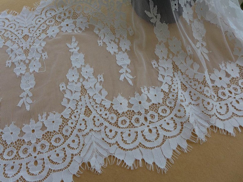 Puede incluir: Borde de encaje blanco con un delicado patr&oacute;n floral y un borde festoneado. El encaje es transparente y tiene una textura suave y fluida.