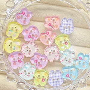 May include: A collection of small, flat, two-hole buttons shaped like rabbit heads in various pastel colors including yellow, pink, blue, and purple. Some buttons feature printed details like small flowers, bows, and gingham patterns on their surfaces.