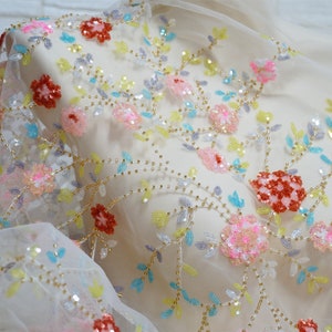 Può includere: Tessuto di tulle bianco con ricamo floreale colorato e paillettes. I fiori sono rosa, rossi, blu e gialli. Il tessuto è perlato con perline dorate.