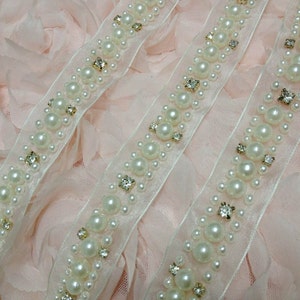 May include: Two white beaded trims with clear rhinestones on a pink fabric background. The trim is made of a sheer fabric with a row of pearls and rhinestones.