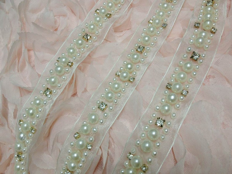 May include: Two white beaded trims with clear rhinestones on a pink fabric background. The trim is made of a sheer fabric with a row of pearls and rhinestones.