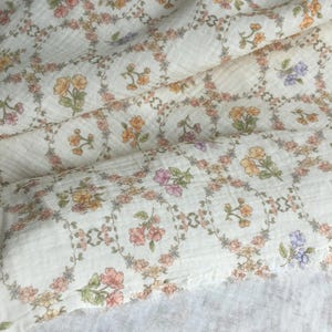 May include: Cream-colored fabric with a floral pattern. The design features small, colorful flowers in shades of pink, orange, and purple, arranged in a repeating square pattern. The fabric has a textured, slightly wrinkled appearance, suggesting a soft material.