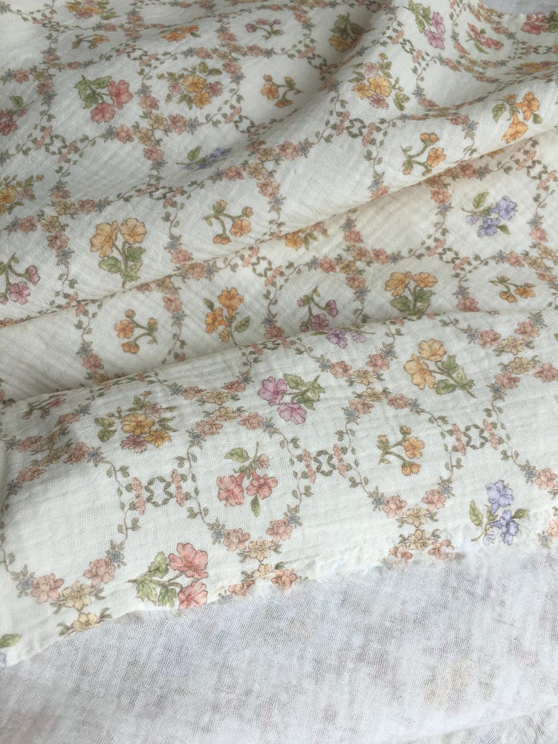 May include: Cream-colored fabric with a floral pattern. The design features small, colorful flowers in shades of pink, orange, and purple, arranged in a repeating square pattern. The fabric has a textured, slightly wrinkled appearance, suggesting a soft material.
