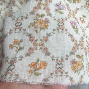 May include: Close-up of a white fabric with a delicate floral pattern. The design features small, colorful flowers in shades of orange, pink, and purple, arranged on a light background. The fabric has a soft, textured appearance.