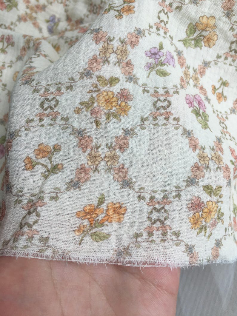 May include: Close-up of a white fabric with a delicate floral pattern. The design features small, colorful flowers in shades of orange, pink, and purple, arranged on a light background. The fabric has a soft, textured appearance.