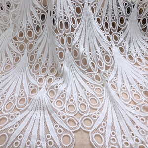 May include: White lace fabric with a detailed peacock feather design. The fabric features a repeating pattern of openwork circles and curved lines, creating a delicate and elegant texture. Ideal for bridal wear or decorative projects.