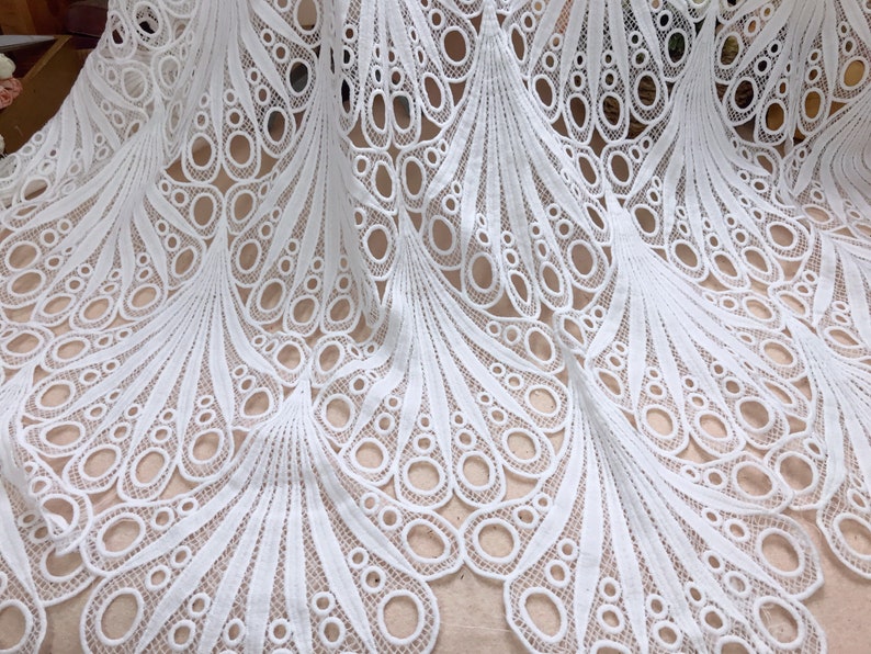 May include: White lace fabric with a detailed peacock feather design. The fabric features a repeating pattern of openwork circles and curved lines, creating a delicate and elegant texture. Ideal for bridal wear or decorative projects.