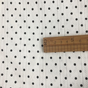 May include: A white fabric featuring a pattern of small black polka dots and vertical textured stripes. A wooden ruler is placed horizontally across the fabric, showing a measurement scale that spans approximately 6 centimeters in length.
