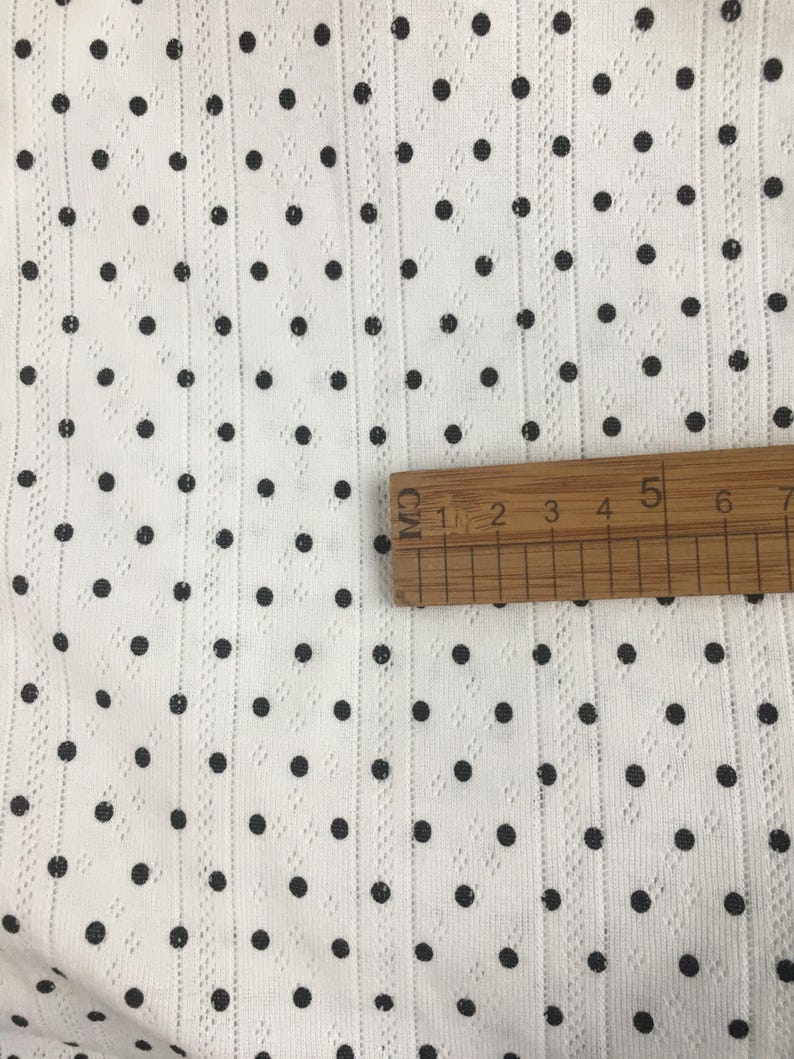 May include: A white fabric featuring a pattern of small black polka dots and vertical textured stripes. A wooden ruler is placed horizontally across the fabric, showing a measurement scale that spans approximately 6 centimeters in length.