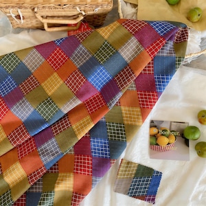 Puede incluir: Una colorida tela de patchwork con un patrón de cuadrícula en tonos de rojo, naranja, amarillo, verde, azul y morado. La tela está doblada, revelando su textura y diseño. También se ve una pequeña muestra de tela.