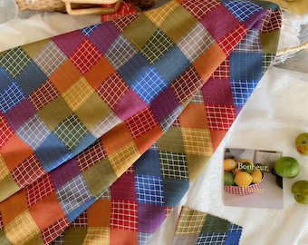 May include: A colorful patchwork fabric with a grid pattern in shades of red, orange, yellow, green, blue, and purple. The fabric is folded, revealing its texture and design. A small swatch of the fabric is also visible.