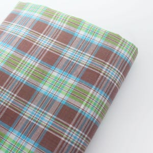 May include: A folded piece of fabric featuring a plaid pattern with intersecting lines in shades of brown, bright lime green, and light sky blue. The textile displays a consistent grid design throughout, with the colours layered to create a woven appearance.