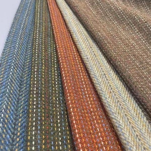 Puede incluir: Una colección de cinco muestras de tela dispuestas en abanico. Cada muestra presenta un color distinto, incluyendo azul, verde oliva, naranja, gris claro y marrón, todas caracterizadas por un tejido de espiga texturizado y rayas verticales.