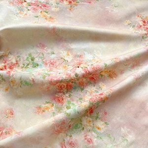 May include: A fabric with a floral pattern featuring pink, peach, and yellow roses with green leaves. The roses are scattered across a light pink and cream-colored background. The fabric is draped, showing folds and texture.