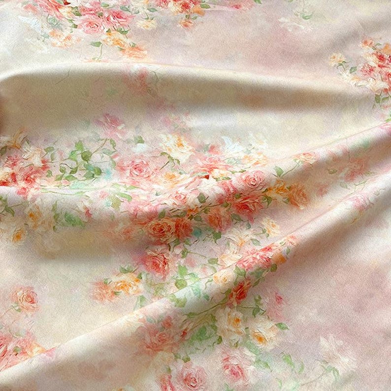 May include: A fabric with a floral pattern featuring pink, peach, and yellow roses with green leaves. The roses are scattered across a light pink and cream-colored background. The fabric is draped, showing folds and texture.