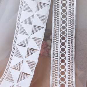 May include: Two white lace trims with different patterns. One trim features a geometric pattern of triangles, while the other has a repeating circle design.