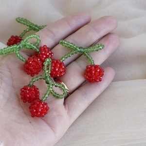 May include: A cluster of beaded cherries with bright red fruit and green stems and leaves. The cherries are made of small, round beads, and the stems and leaves are made of smaller, green beads. The cherries are a handmade craft item.