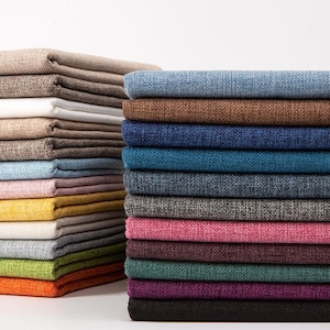 Op de afbeelding: Stapels gevouwen stof in diverse kleuren, waaronder beige, wit, blauw, bruin, teal, grijs, roze en paars. De stof heeft een getextureerde uitstraling, wat wijst op linnen of een vergelijkbaar materiaal. De afbeelding toont een scala aan kleuren, geschikt voor bekleding of textielprojecten.