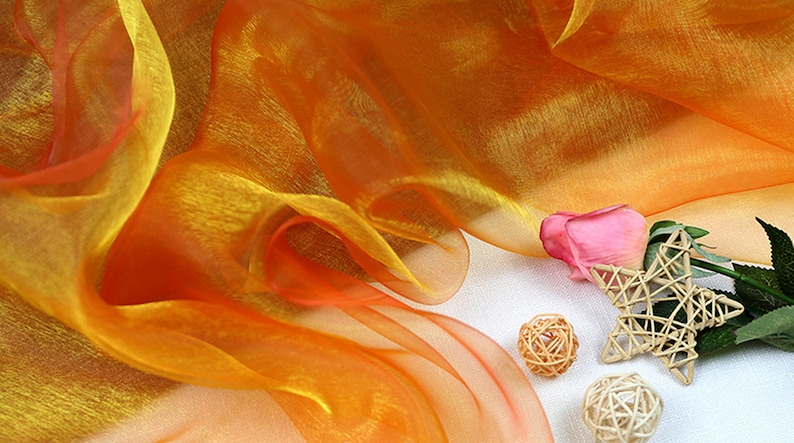May include: Orange and yellow sheer fabric with a pink rose and a woven star on a white background.