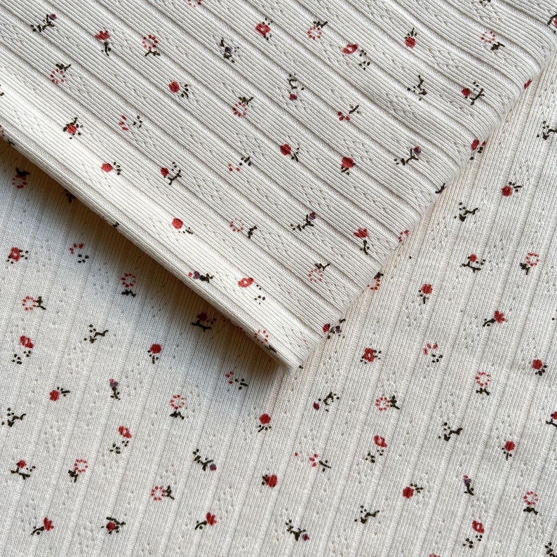 May include: Close-up of a white ribbed fabric with a delicate floral pattern. Small red and purple flowers are scattered across the surface, creating a charming and feminine design. The fabric has a textured, ribbed appearance.