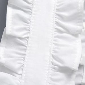 May include: Close-up of a white fabric detail with layered ruffles. The ruffles are neatly sewn with visible stitching, creating a textured effect. The fabric appears to be crisp and clean, with a smooth, slightly shiny finish.