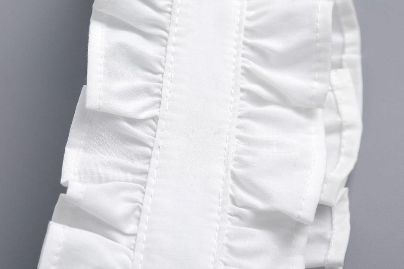 May include: Close-up of a white fabric detail with layered ruffles. The ruffles are neatly sewn with visible stitching, creating a textured effect. The fabric appears to be crisp and clean, with a smooth, slightly shiny finish.
