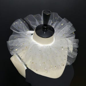 Pode incluir: Um colarinho de tule branco com um padrão de pequenas estrelas e luas crescentes douradas. O colarinho é construído com várias camadas de tecido transparente franzido que criam uma forma circular volumosa ao redor do pescoço.