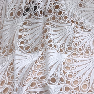 May include: Close-up of white lace fabric with a detailed peacock feather design. The fabric features a repeating pattern of open circles and curved lines, creating a delicate and airy texture. The lace is layered, showing the intricate design.