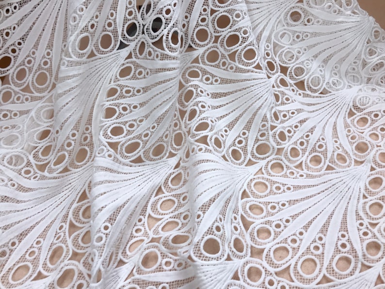 May include: Close-up of white lace fabric with a detailed peacock feather design. The fabric features a repeating pattern of open circles and curved lines, creating a delicate and airy texture. The lace is layered, showing the intricate design.