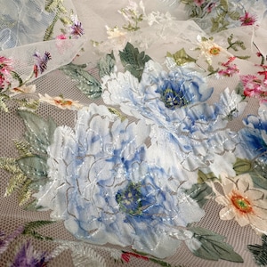 Puede incluir: Tela blanca transparente con dise&ntilde;os florales bordados en azul y verde. La tela tiene una textura de malla delicada y presenta grandes flores azules con detalles intrincados.