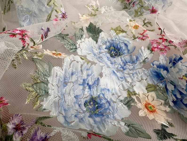 Puede incluir: Tela blanca transparente con dise&ntilde;os florales bordados en azul y verde. La tela tiene una textura de malla delicada y presenta grandes flores azules con detalles intrincados.