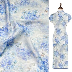 May include: A sleeveless cheongsam dress featuring a high mandarin collar and a fitted silhouette. The fabric is white with a repeating pattern of blue hydrangea flowers and soft green leaves, displayed both as draped fabric and on a dress form.