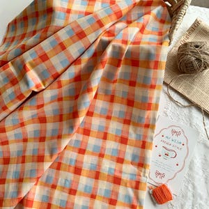 Op de afbeelding: Een gevouwen stof met een geruit patroon in oranje, rood, geel en lichtblauw. De stof is gedrapeerd over een wit oppervlak, met een mand, touw en een kaart met de tekst "My Wish French Style".