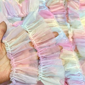 May include: A length of sheer, ruffled fabric trim featuring a soft, multi-coloured pastel gradient. The translucent material is gathered into dense, wavy pleats, displaying alternating shades of light pink, pale blue, yellow, and lavender throughout.