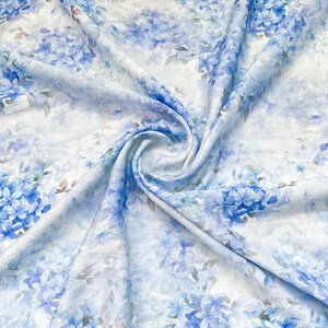 May include: A lightweight fabric featuring a repeating pattern of blue hydrangea flowers on a white background. The material is gathered in the centre, creating soft folds and drapes that highlight the delicate floral print and texture.