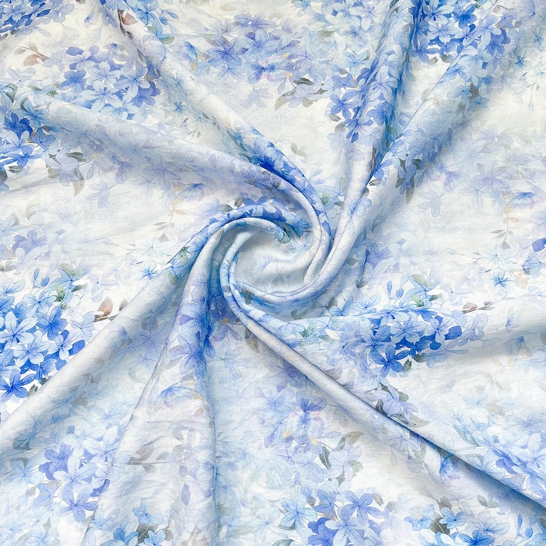 May include: A lightweight fabric featuring a repeating pattern of blue hydrangea flowers on a white background. The material is gathered in the centre, creating soft folds and drapes that highlight the delicate floral print and texture.
