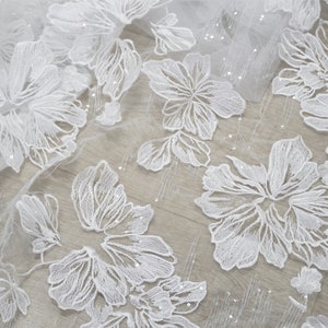 May include: White floral lace fabric with a delicate pattern and a subtle shimmer. The fabric is sheer and lightweight, making it ideal for a variety of sewing projects.
