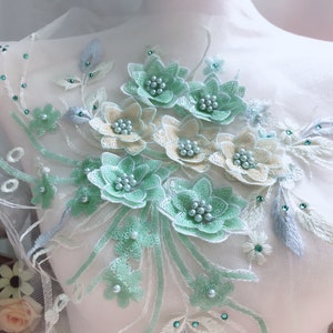 May include: A delicate floral applique with green, white, and blue flowers and leaves. The applique is made of lace and sequins and features pearl accents.