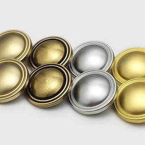 Può includere: Una collezione di otto pomelli per mobili in varie finiture metalliche. I pomelli sono rotondi con un centro leggermente bombato e un bordo esterno decorativo. Le finiture includono oro, ottone anticato e argento.