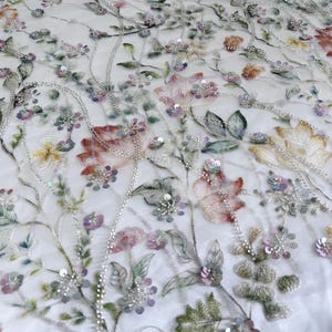 Könnte beinhalten: Nahaufnahme eines weißen Stoffes mit gestickten Blumenmustern. Das Design zeigt Blumen in Rosa, Pfirsich, Gelb und Lila, mit grünen Blättern und silbernen Perlenakzenten. Der Stoff eignet sich für Kleidung oder Heimdekoration.