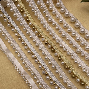 May include: Close-up of various decorative trims featuring white faux pearls and gold-coloured beads. The trims are arranged diagonally, showcasing different textures and designs. Some trims have a satin ribbon base, while others have a woven fabric base.
