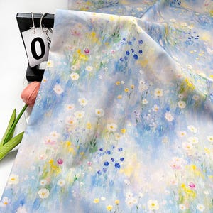 Puede incluir: Tela floral con un diseño estilo acuarela. La tela presenta un fondo azul claro con flores blancas, amarillas, rosas y azules. La tela está drapeada, mostrando el flujo del patrón. Un expositor negro con un letrero "0" y un tulipán rosa están en el fondo.