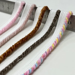 May include: Five strips of velvet piping trim aligned against a metal ruler. The colors include light pink, a darker pink, brown, dark gray, and a multi-colored pastel twist. Each strip features a white woven fabric edge for sewing attachment.