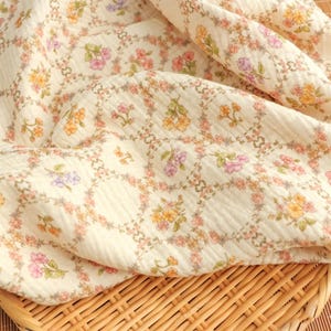 May include: Cream-colored fabric with a floral pattern. The design features small flowers in shades of pink, yellow, and purple, arranged in a repeating pattern. The fabric has a textured, slightly wrinkled appearance.