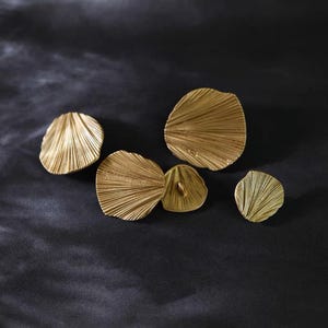 May include: Five gold-coloured, fan-shaped decorative objects with a textured, ridged surface. The objects are arranged on a dark, textured background. The shapes vary in size, with the largest being approximately 7.6 cm in diameter.