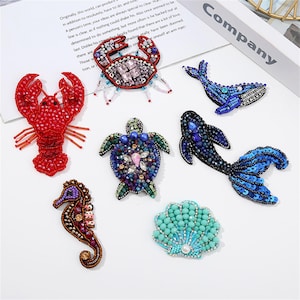 May include: A selection of beaded brooches in the shape of marine creatures: a red lobster, a crab, a whale, a sea turtle, a seahorse, a fish, and a seashell. The brooches are adorned with colourful beads and sequins. The word "Company" is visible in the background.