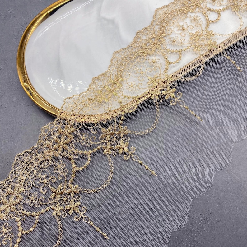 May include: Gold lace trim with a delicate floral pattern and a scalloped edge. The lace is on a sheer mesh background.